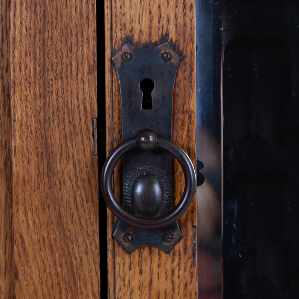 An Arts and Crafts oak hall wardrobe featuring a central mirrored door, dark glass roundel inserts near the top, and an arched apron base. View XI