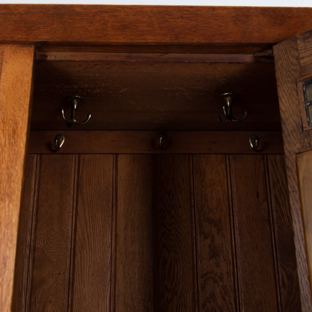 An Arts and Crafts oak hall wardrobe featuring a central mirrored door, dark glass roundel inserts near the top, and an arched apron base. View XII