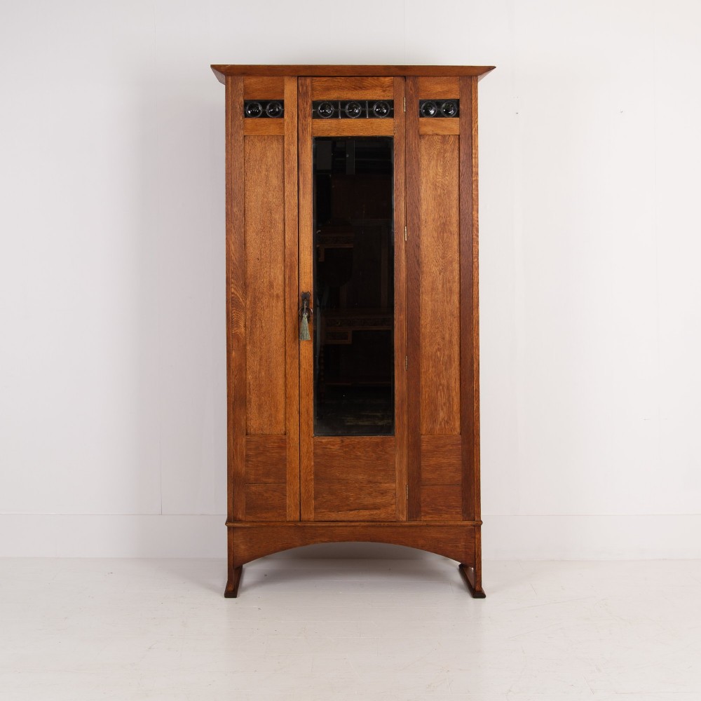 An Arts and Crafts oak hall wardrobe featuring a central mirrored door, dark glass roundel inserts near the top, and an arched apron base. View XV