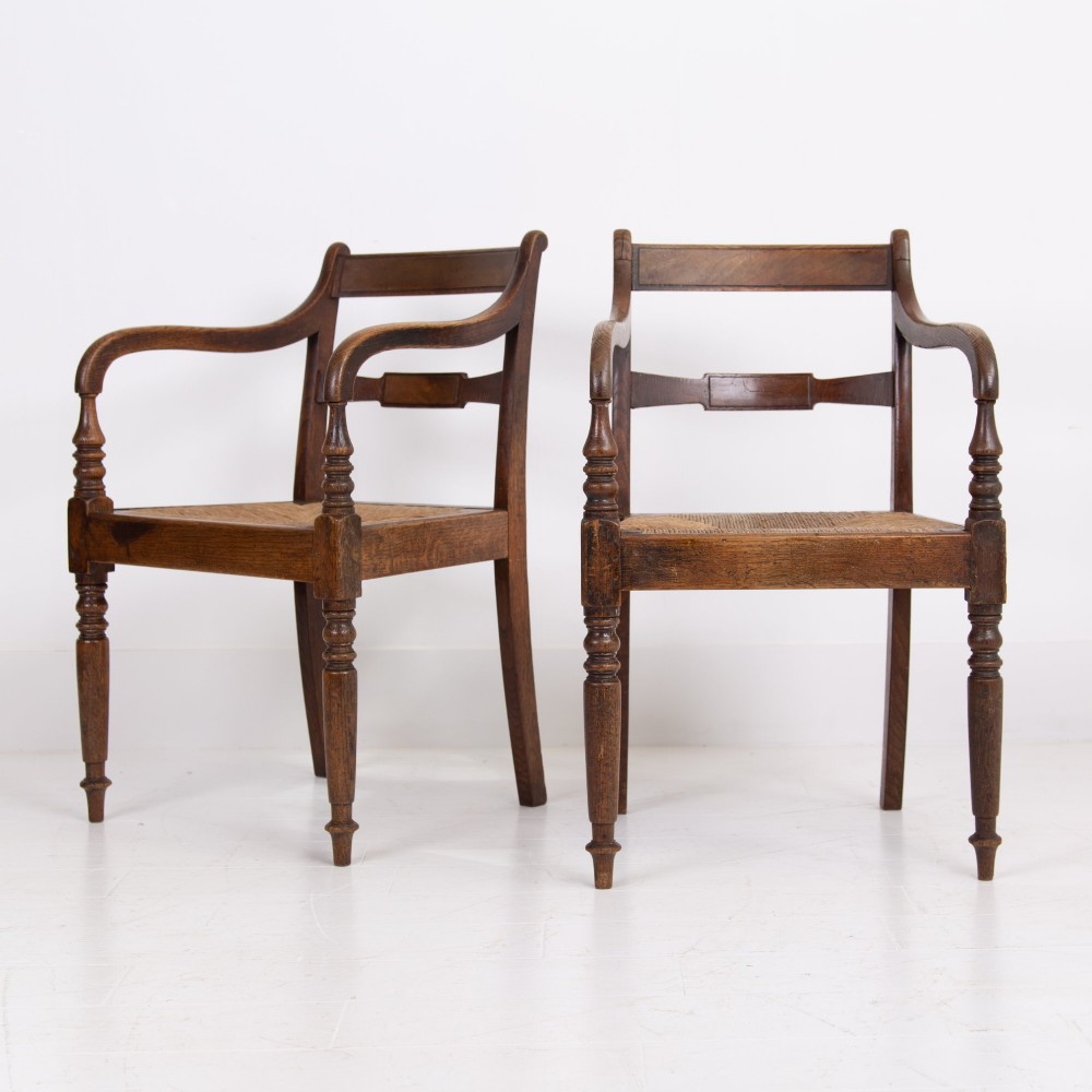 A pair of 1820s Georgian dark oak elbow chairs featuring woven rush seats, swept armrests, and turned front legs.