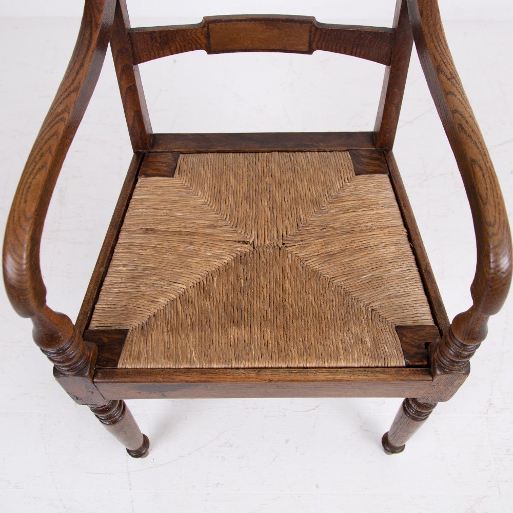 A pair of 1820s Georgian dark oak elbow chairs featuring woven rush seats, swept armrests, and turned front legs. View X