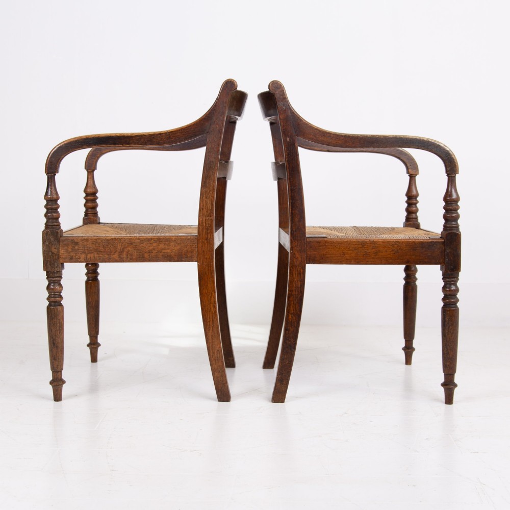 A pair of 1820s Georgian dark oak elbow chairs featuring woven rush seats, swept armrests, and turned front legs. View XIV