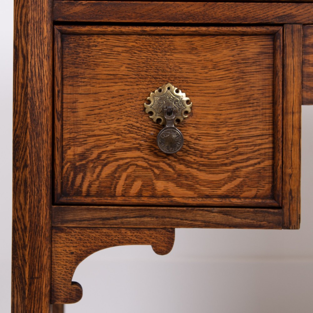 A golden oak writing desk featuring four drawers with brass drop handles, shaped kneehole brackets, turned legs, and metal castors. View V