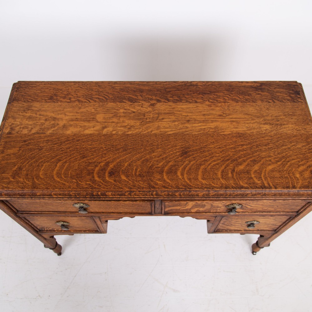 A golden oak writing desk featuring four drawers with brass drop handles, shaped kneehole brackets, turned legs, and metal castors. View VI