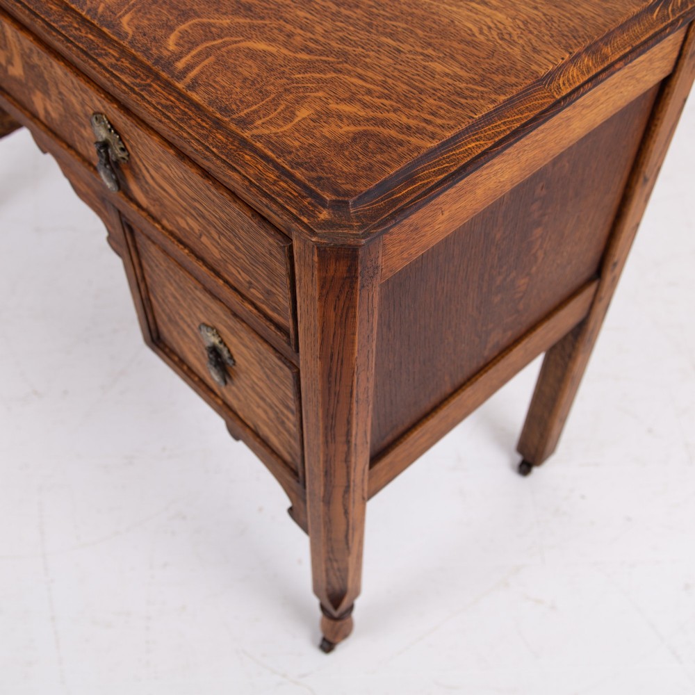 A golden oak writing desk featuring four drawers with brass drop handles, shaped kneehole brackets, turned legs, and metal castors. View VIII