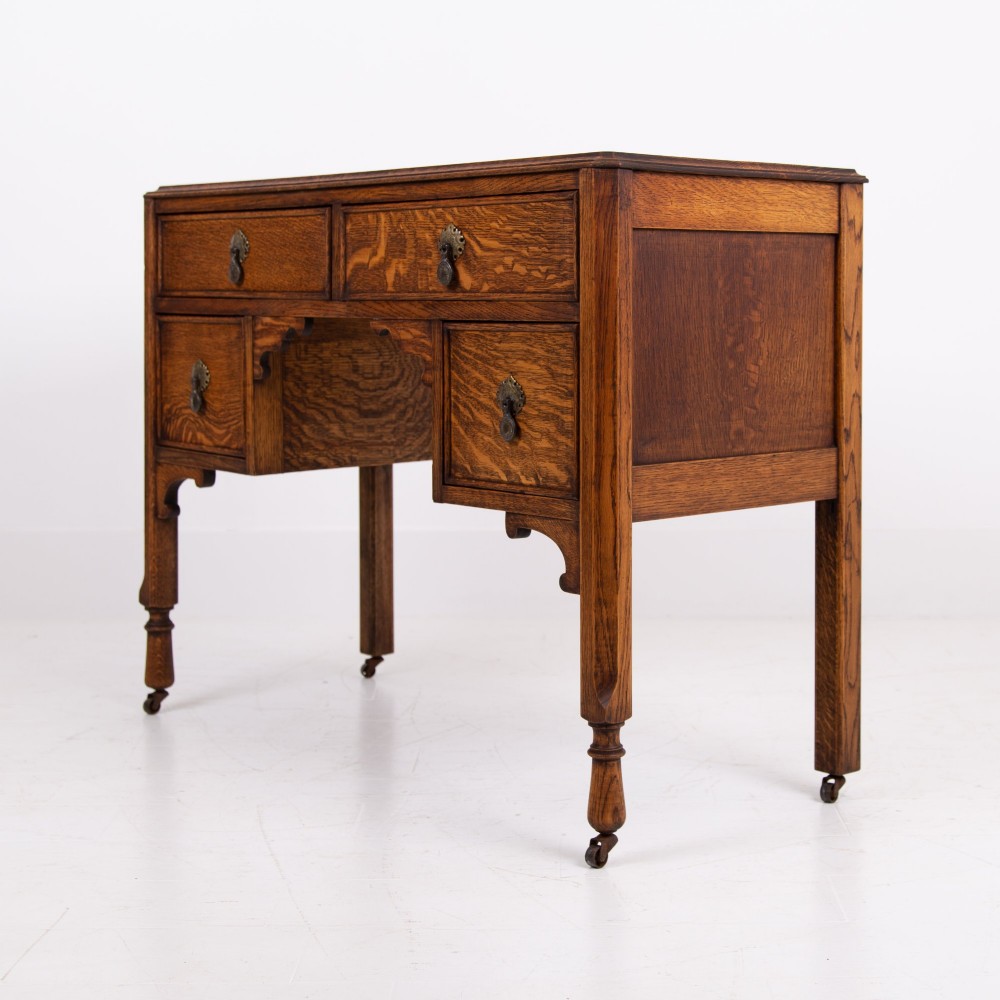 A golden oak writing desk featuring four drawers with brass drop handles, shaped kneehole brackets, turned legs, and metal castors. View IX