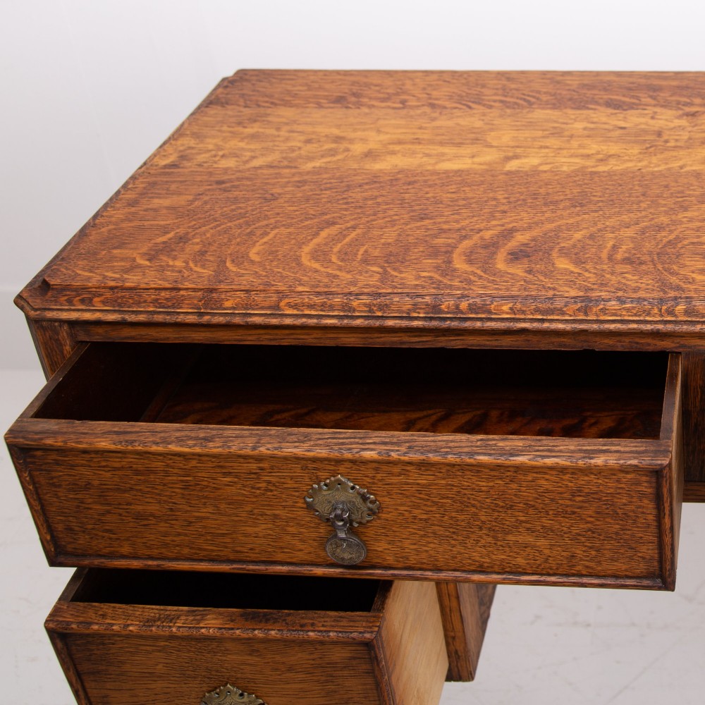 A golden oak writing desk featuring four drawers with brass drop handles, shaped kneehole brackets, turned legs, and metal castors. View X