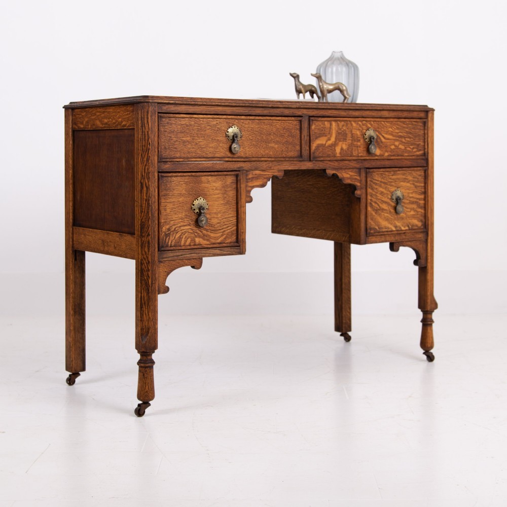 A golden oak writing desk featuring four drawers with brass drop handles, shaped kneehole brackets, turned legs, and metal castors. View XIV