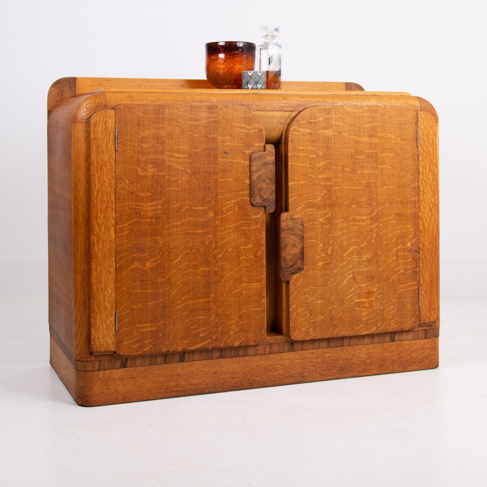 An Art Deco golden oak sideboard cabinet featuring rounded top edges, quarter-sawn wood grain, two central doors with wooden block handles, and a solid base plinth.