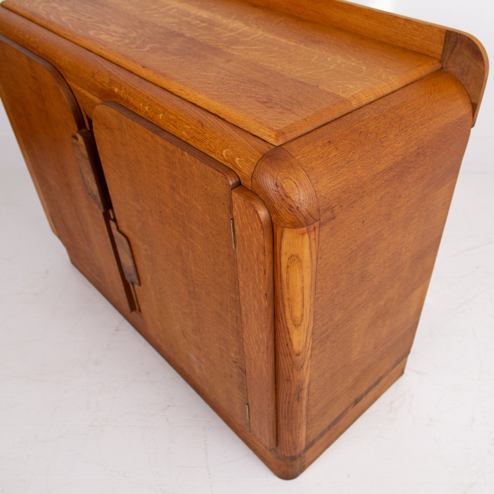 An Art Deco golden oak sideboard cabinet featuring rounded top edges, quarter-sawn wood grain, two central doors with wooden block handles, and a solid base plinth. View IV