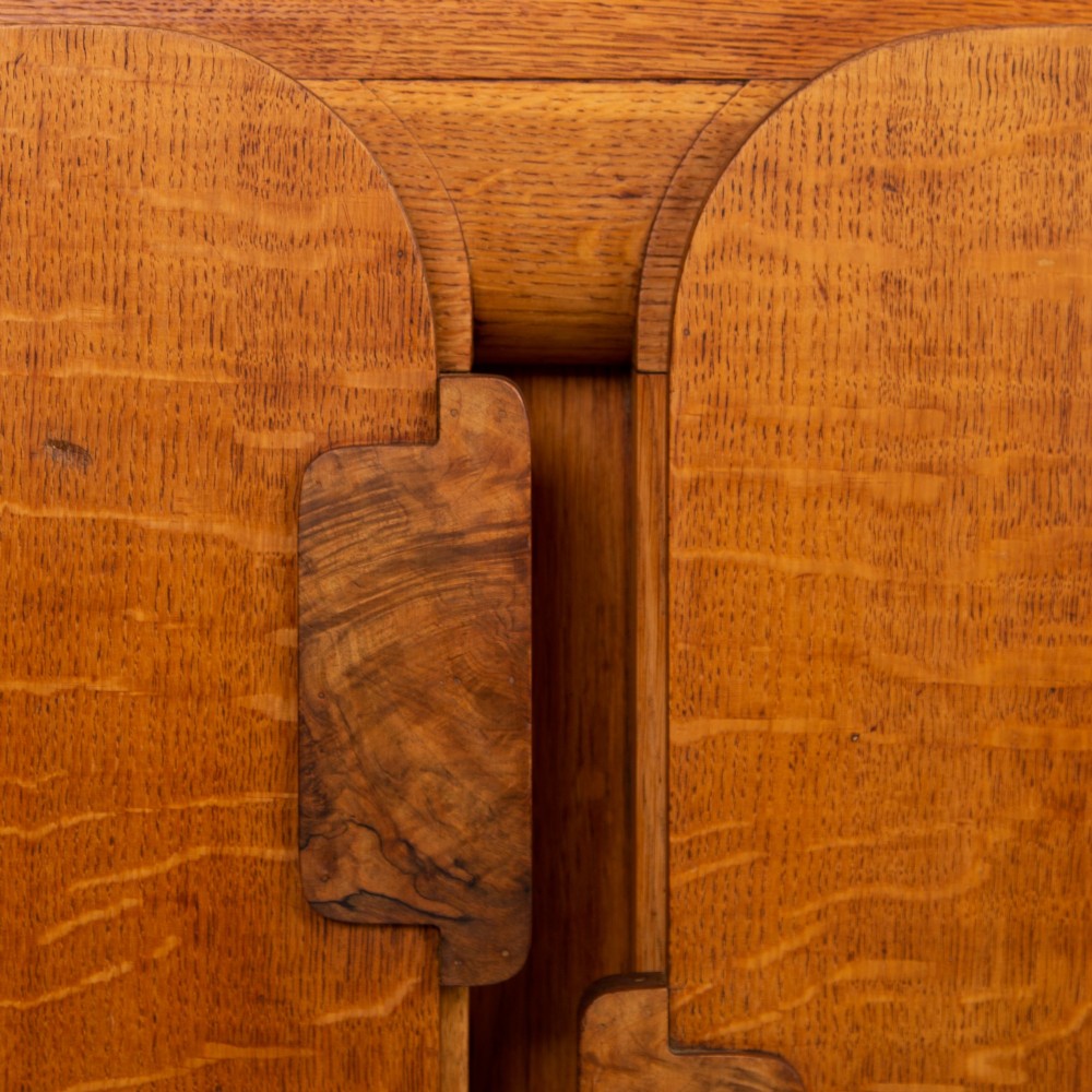 An Art Deco golden oak sideboard cabinet featuring rounded top edges, quarter-sawn wood grain, two central doors with wooden block handles, and a solid base plinth. View VI