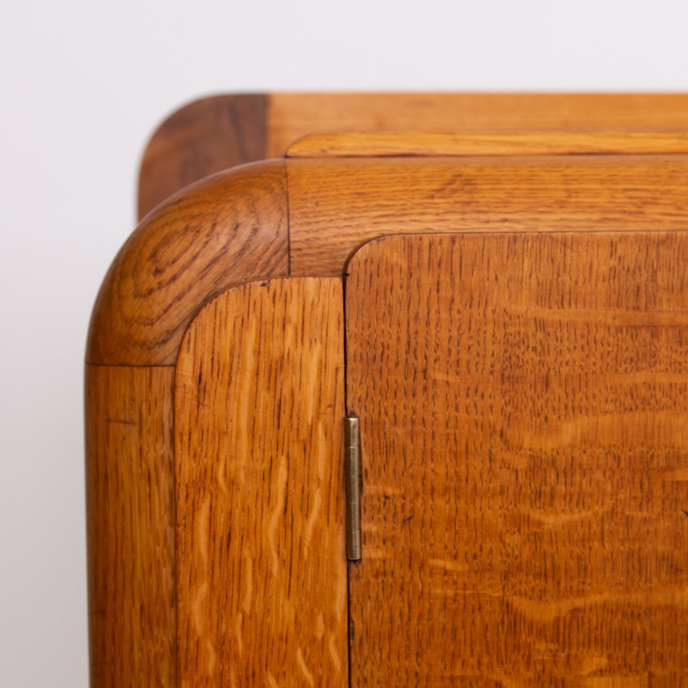 An Art Deco golden oak sideboard cabinet featuring rounded top edges, quarter-sawn wood grain, two central doors with wooden block handles, and a solid base plinth. View VII