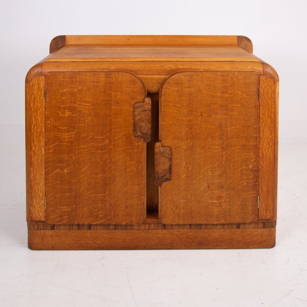 An Art Deco golden oak sideboard cabinet featuring rounded top edges, quarter-sawn wood grain, two central doors with wooden block handles, and a solid base plinth. View X