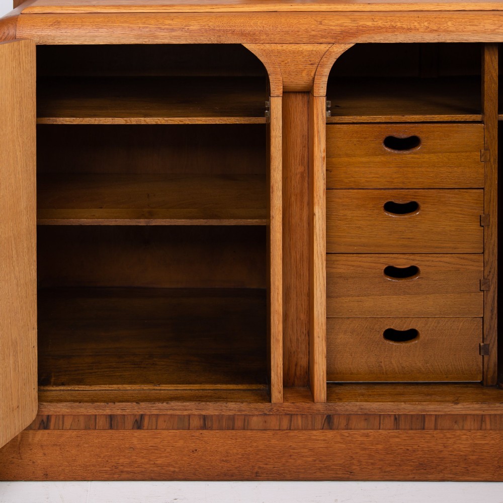 An Art Deco golden oak sideboard cabinet featuring rounded top edges, quarter-sawn wood grain, two central doors with wooden block handles, and a solid base plinth. View XI