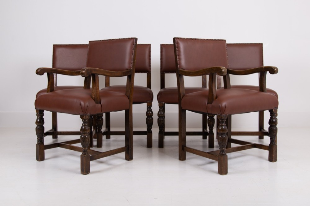 A set of six dark oak dining chairs featuring brown leather upholstered seats and backrests with brass stud detailing and turned front legs.