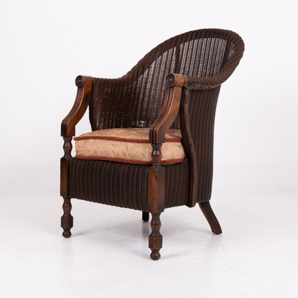 A 1930s dark brown Lloyd Loom woven armchair featuring carved oak armrests, turned front legs, and a patterned seat cushion. View II