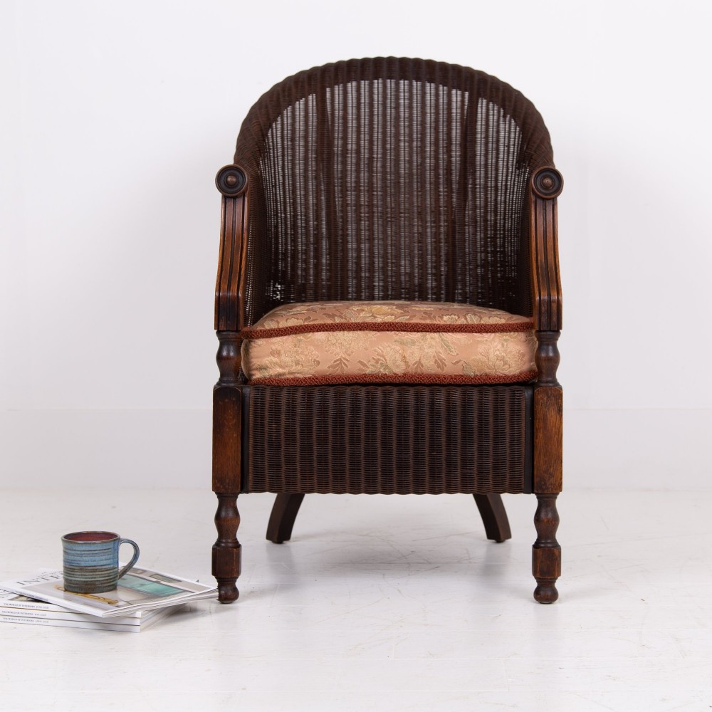 A 1930s dark brown Lloyd Loom woven armchair featuring carved oak armrests, turned front legs, and a patterned seat cushion. View IV