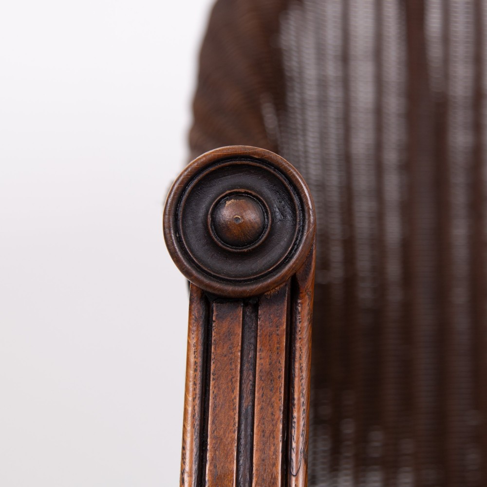 A 1930s dark brown Lloyd Loom woven armchair featuring carved oak armrests, turned front legs, and a patterned seat cushion. View V