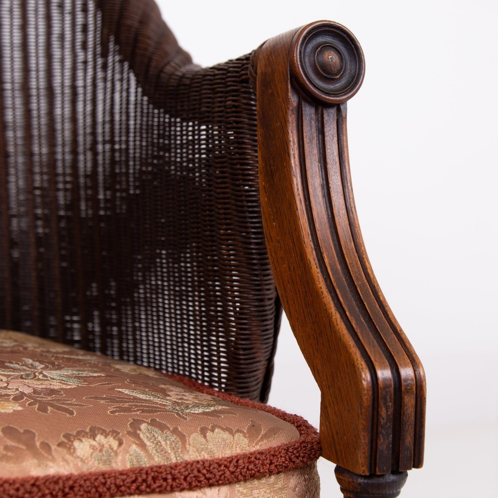 A 1930s dark brown Lloyd Loom woven armchair featuring carved oak armrests, turned front legs, and a patterned seat cushion. View VI