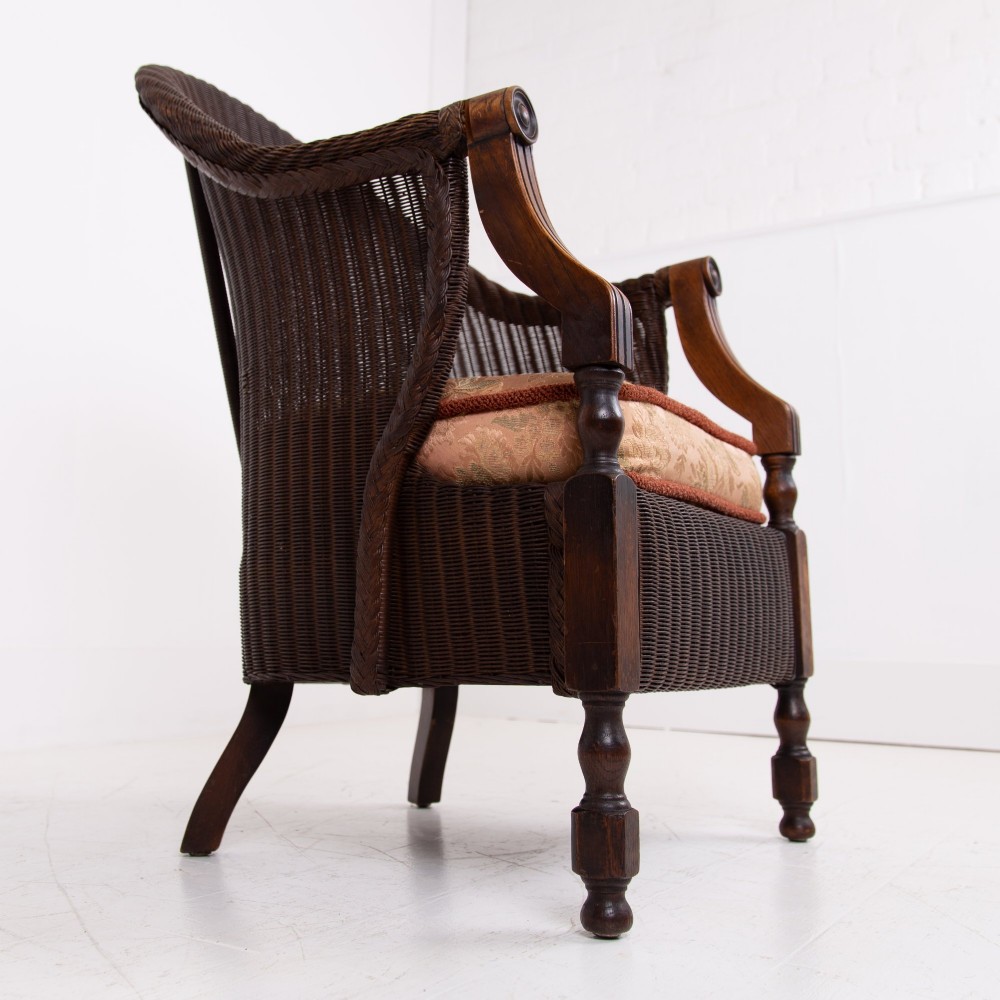 A 1930s dark brown Lloyd Loom woven armchair featuring carved oak armrests, turned front legs, and a patterned seat cushion. View VII