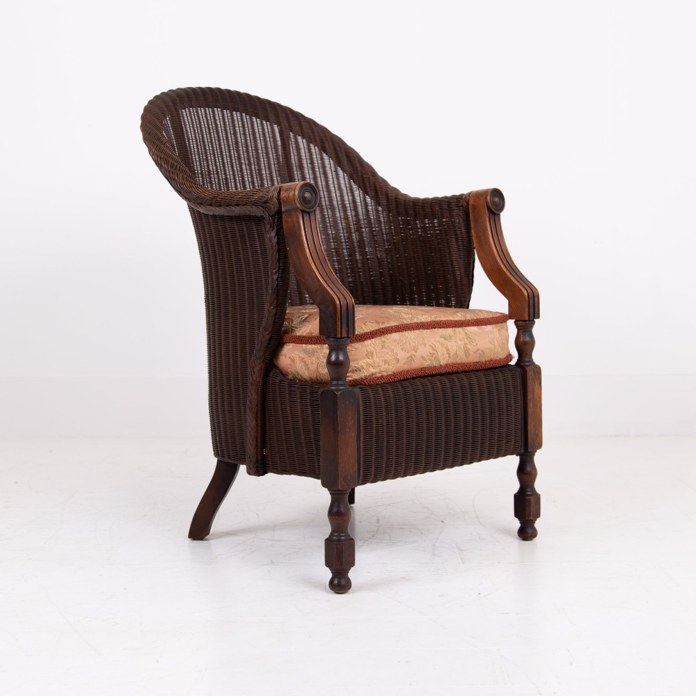 A 1930s dark brown Lloyd Loom woven armchair featuring carved oak armrests, turned front legs, and a patterned seat cushion. View IX