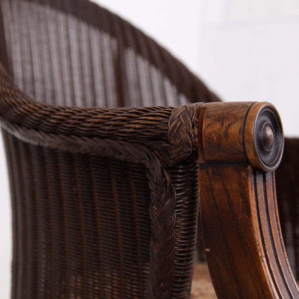 A 1930s dark brown Lloyd Loom woven armchair featuring carved oak armrests, turned front legs, and a patterned seat cushion. View X