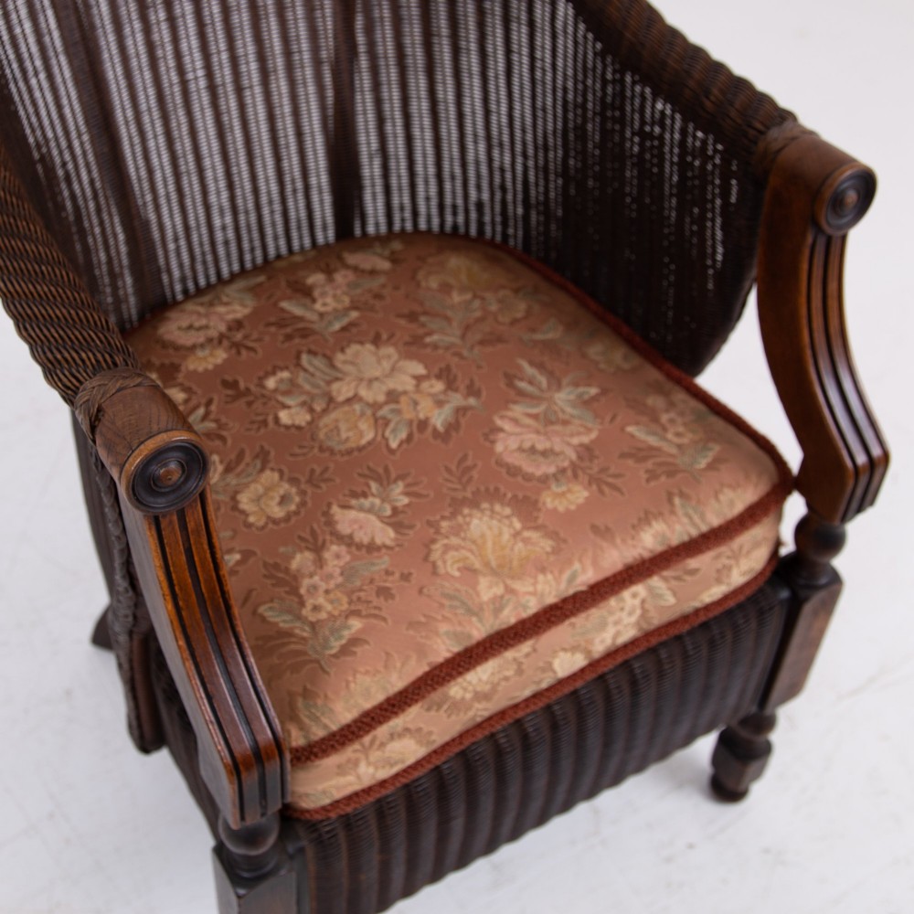 A 1930s dark brown Lloyd Loom woven armchair featuring carved oak armrests, turned front legs, and a patterned seat cushion. View XII