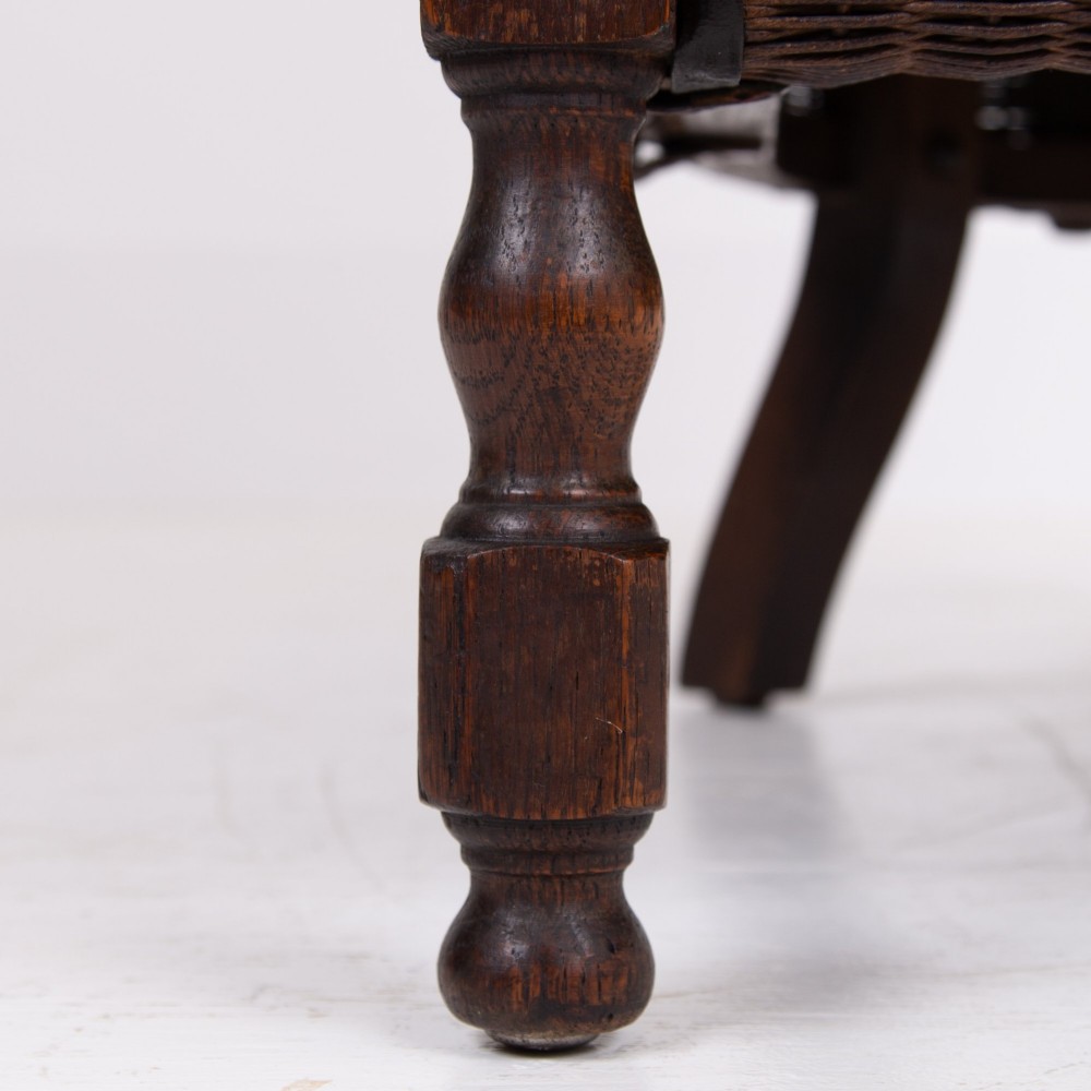 A 1930s dark brown Lloyd Loom woven armchair featuring carved oak armrests, turned front legs, and a patterned seat cushion. View XIII