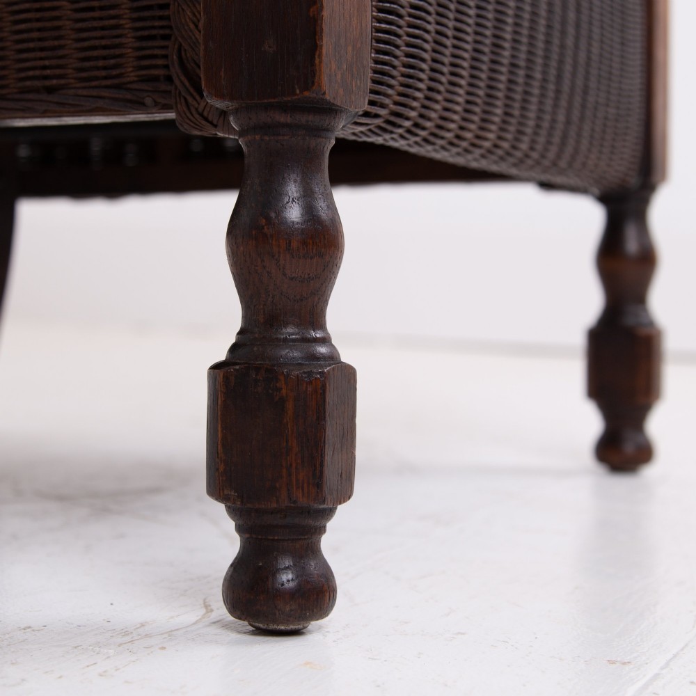 A 1930s dark brown Lloyd Loom woven armchair featuring carved oak armrests, turned front legs, and a patterned seat cushion. View XIV