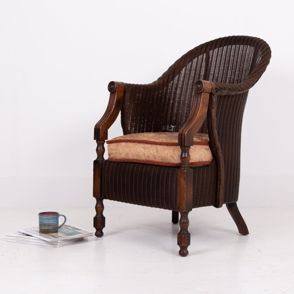 A 1930s dark brown Lloyd Loom woven armchair featuring carved oak armrests, turned front legs, and a patterned seat cushion. View XV