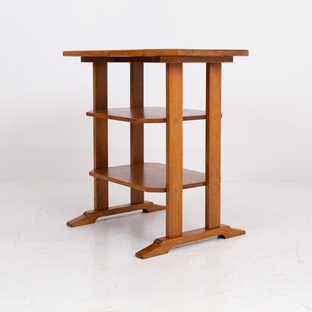 An antique Cotswold School solid oak side table featuring three rectangular shelves supported by straight uprights and sled feet.