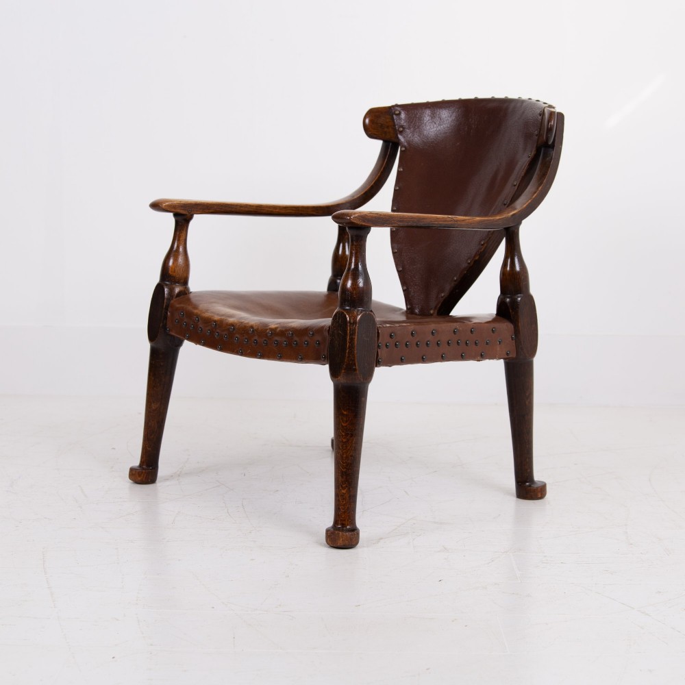 An Arts and Crafts style dark oak armchair featuring turned front legs, wide curved armrests, and brown studded leather upholstery. View III