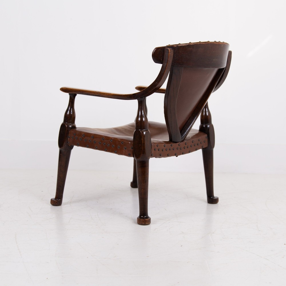 An Arts and Crafts style dark oak armchair featuring turned front legs, wide curved armrests, and brown studded leather upholstery. View IV