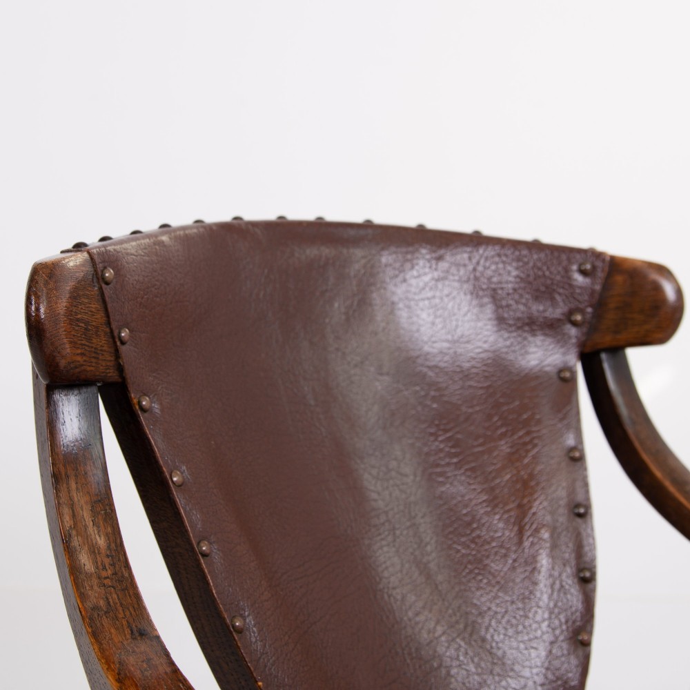 An Arts and Crafts style dark oak armchair featuring turned front legs, wide curved armrests, and brown studded leather upholstery. View V