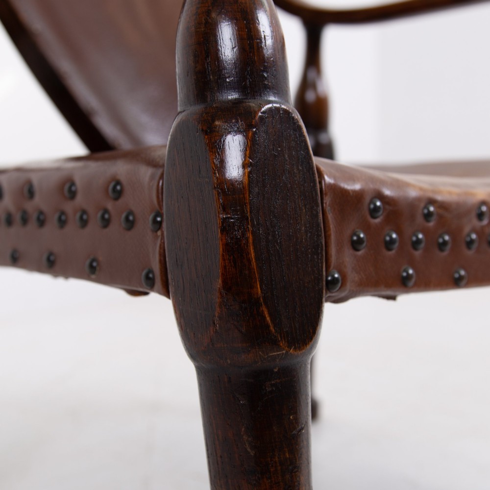 An Arts and Crafts style dark oak armchair featuring turned front legs, wide curved armrests, and brown studded leather upholstery. View VI