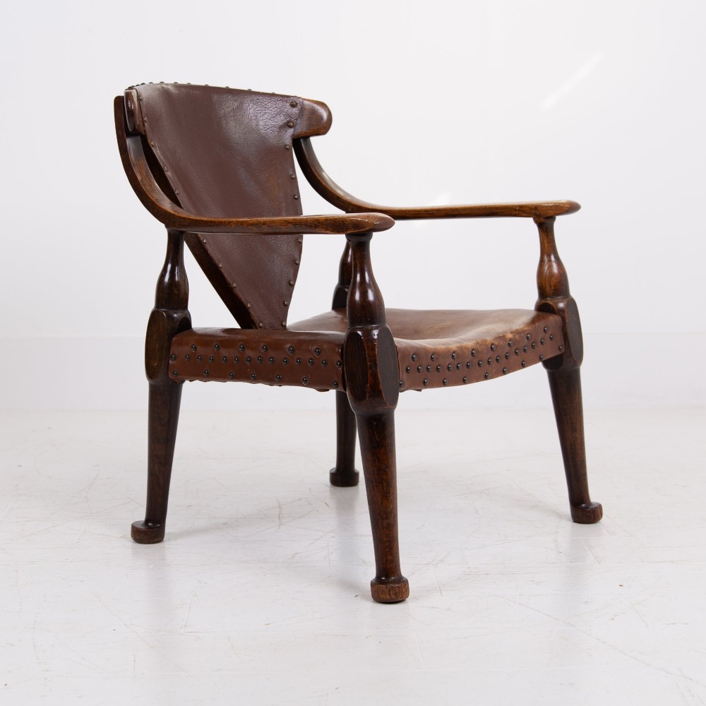 An Arts and Crafts style dark oak armchair featuring turned front legs, wide curved armrests, and brown studded leather upholstery. View VIII