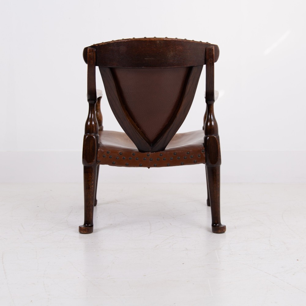 An Arts and Crafts style dark oak armchair featuring turned front legs, wide curved armrests, and brown studded leather upholstery. View IX