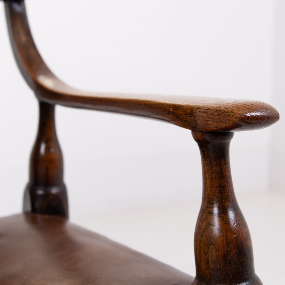 An Arts and Crafts style dark oak armchair featuring turned front legs, wide curved armrests, and brown studded leather upholstery. View X