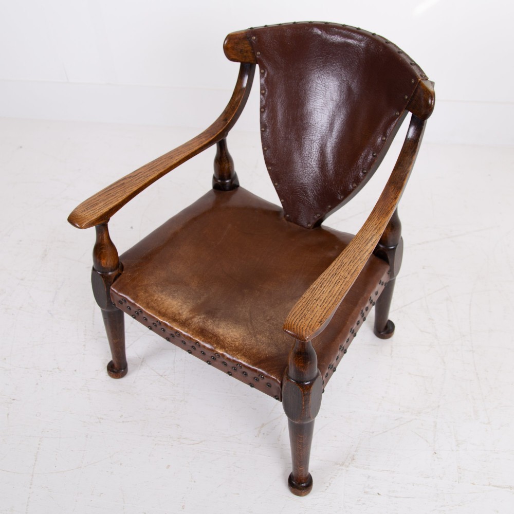 An Arts and Crafts style dark oak armchair featuring turned front legs, wide curved armrests, and brown studded leather upholstery. View XII