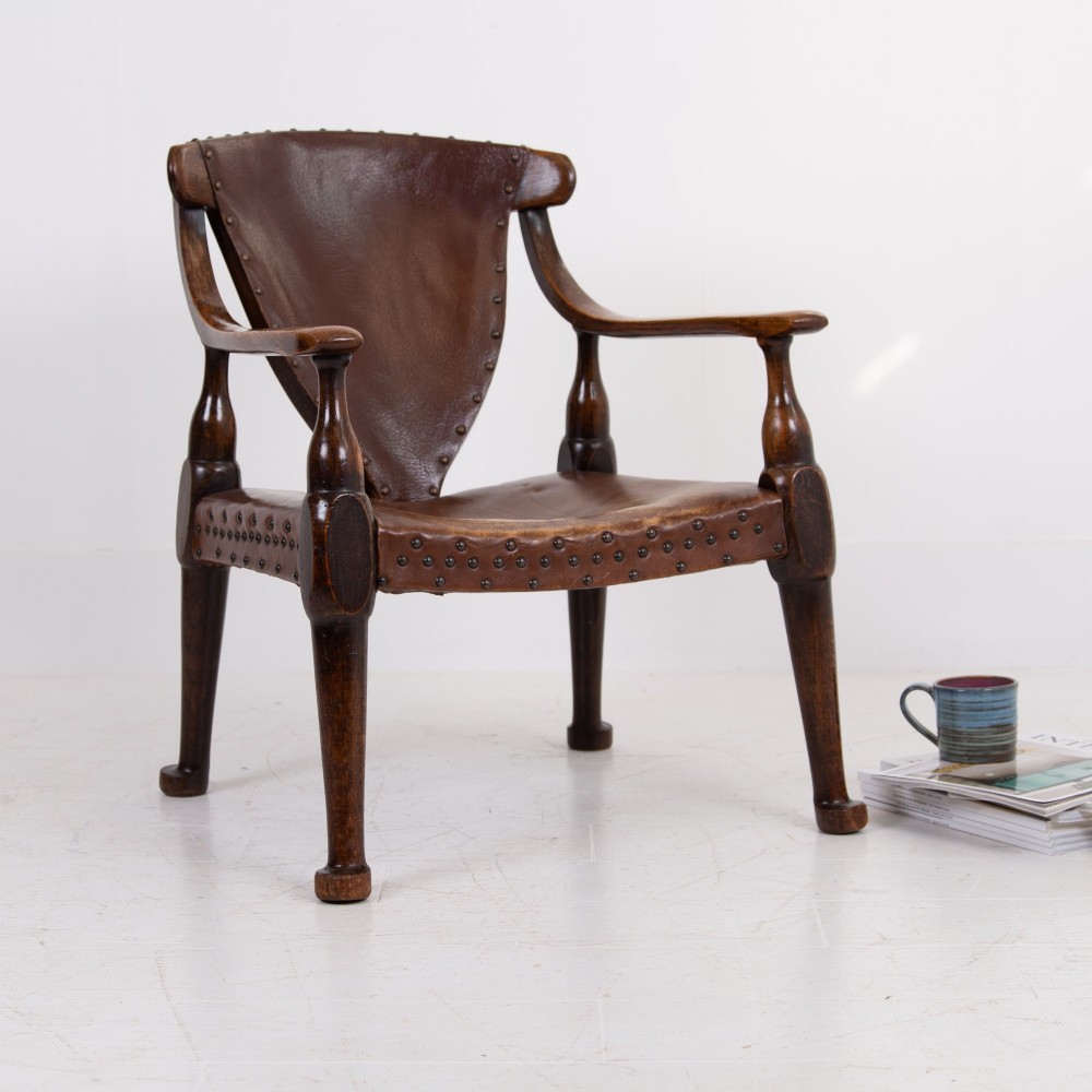 An Arts and Crafts style dark oak armchair featuring turned front legs, wide curved armrests, and brown studded leather upholstery. View XIV
