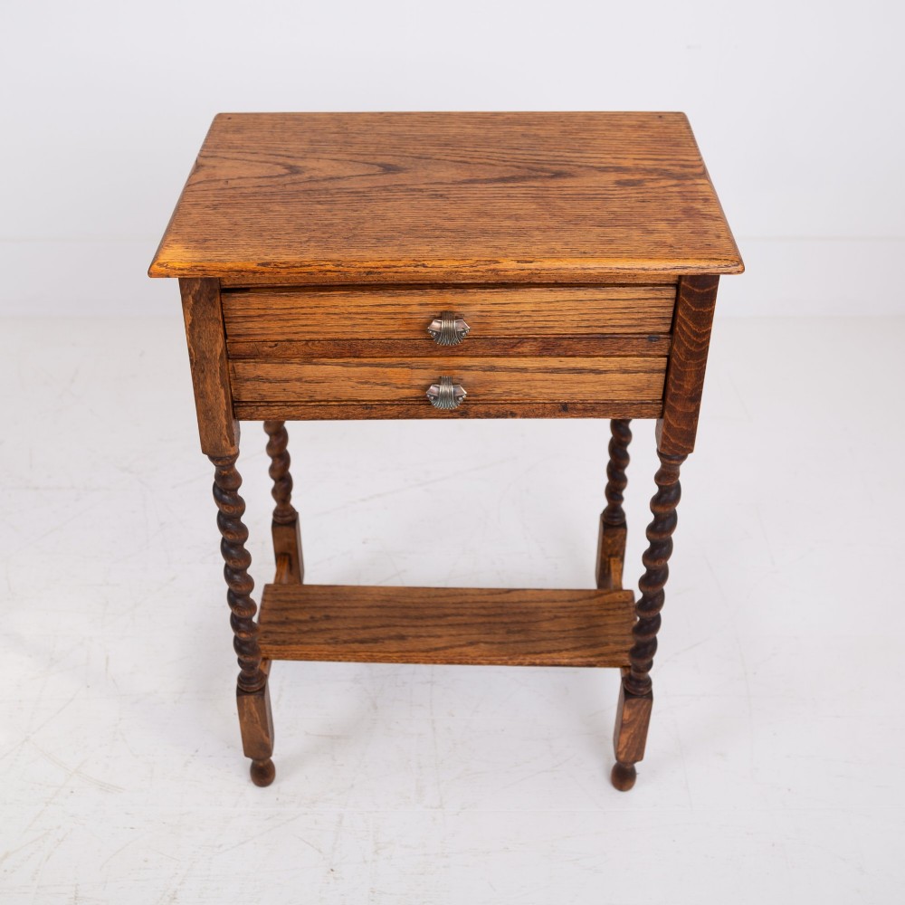 A warm brown oak side table featuring two stacked drawers with metal shell handles, four turned barley twist legs, and a low rectangular shelf.