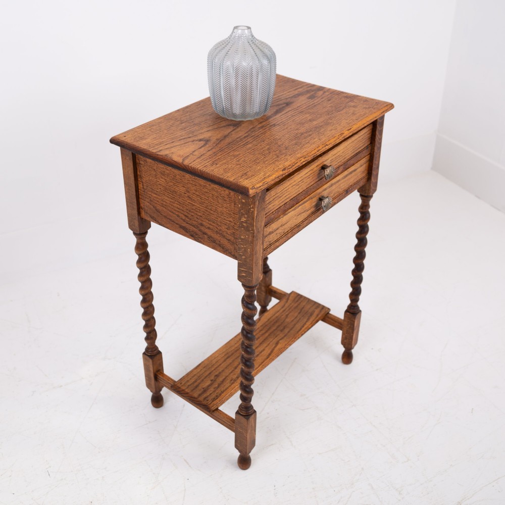 A warm brown oak side table featuring two stacked drawers with metal shell handles, four turned barley twist legs, and a low rectangular shelf. View III