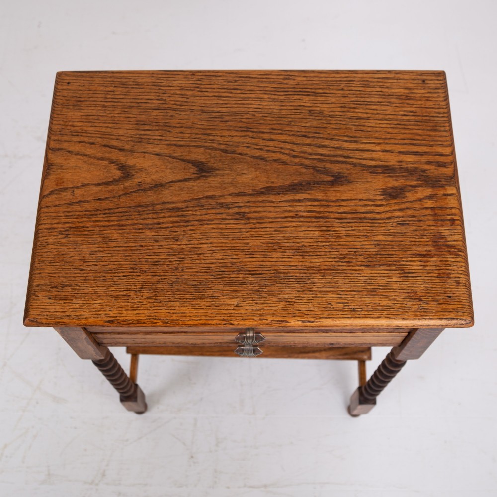 A warm brown oak side table featuring two stacked drawers with metal shell handles, four turned barley twist legs, and a low rectangular shelf. View VI