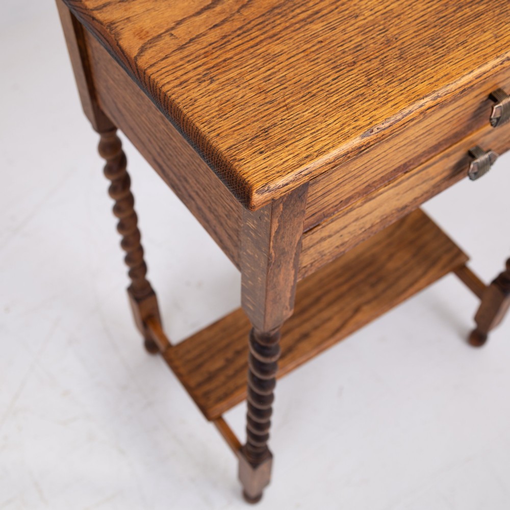 A warm brown oak side table featuring two stacked drawers with metal shell handles, four turned barley twist legs, and a low rectangular shelf. View VII