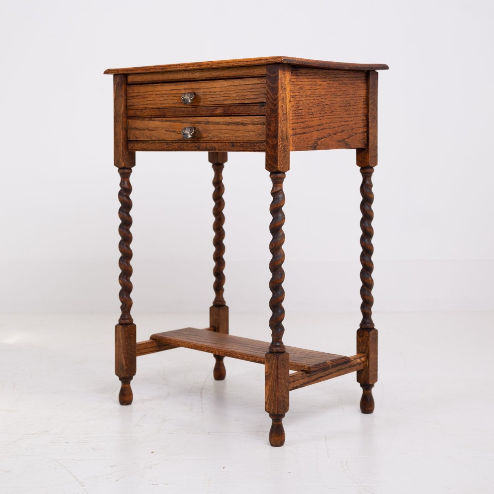 A warm brown oak side table featuring two stacked drawers with metal shell handles, four turned barley twist legs, and a low rectangular shelf. View IX