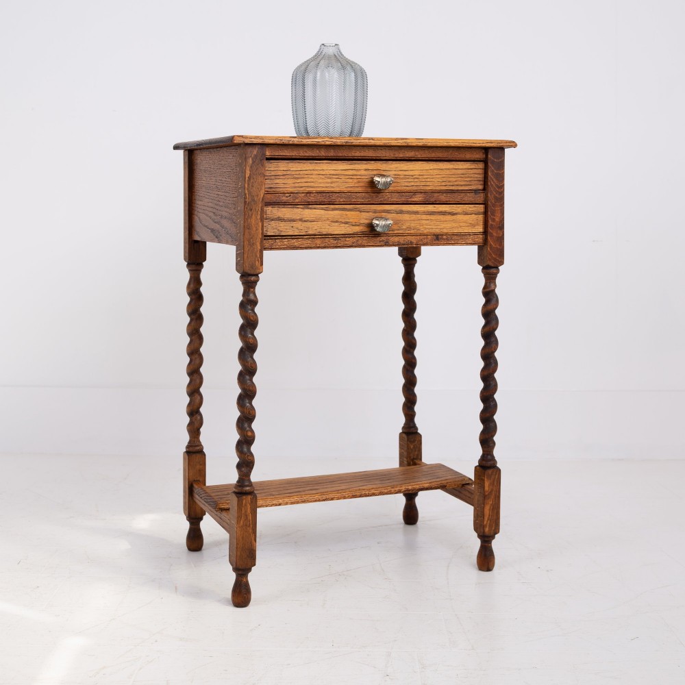 A warm brown oak side table featuring two stacked drawers with metal shell handles, four turned barley twist legs, and a low rectangular shelf. View XI
