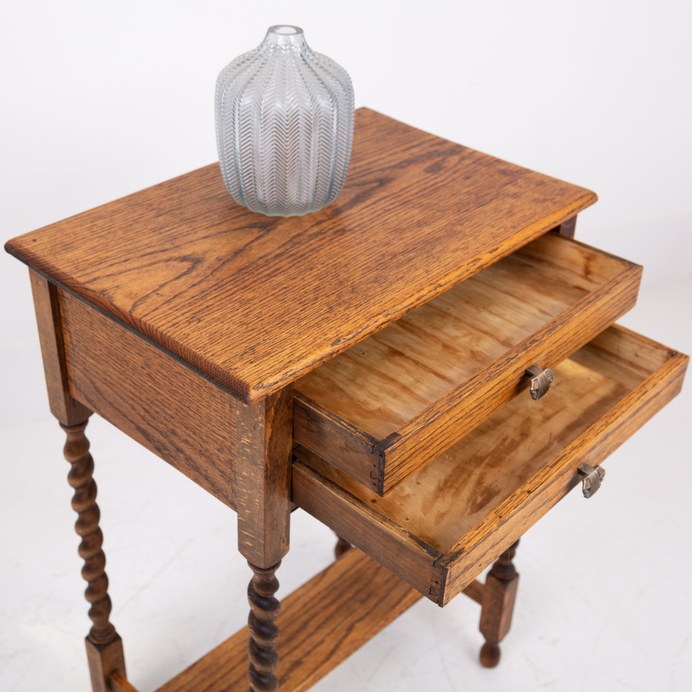 A warm brown oak side table featuring two stacked drawers with metal shell handles, four turned barley twist legs, and a low rectangular shelf. View XII