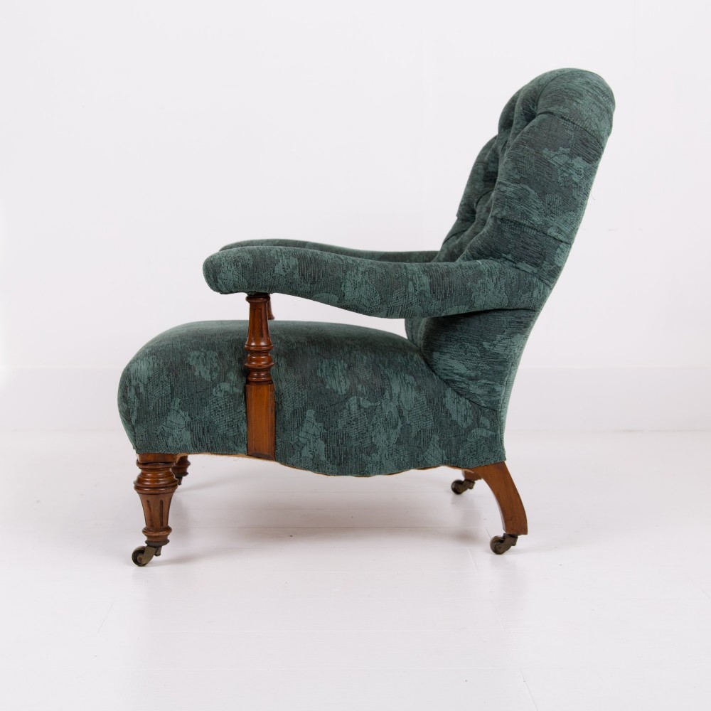 A profile view of a Victorian walnut open library armchair with dark green button-backed upholstery, turned front legs, and brass castors.