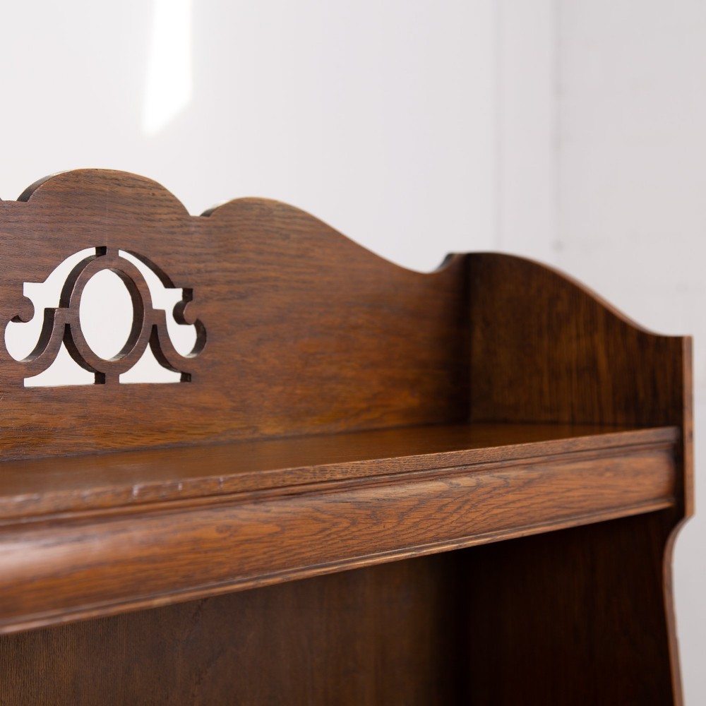 An Arts and Crafts solid oak open bookcase featuring five shelves, shaped side panels, and a decorative cresting rail with a pierced central cutout. View VII