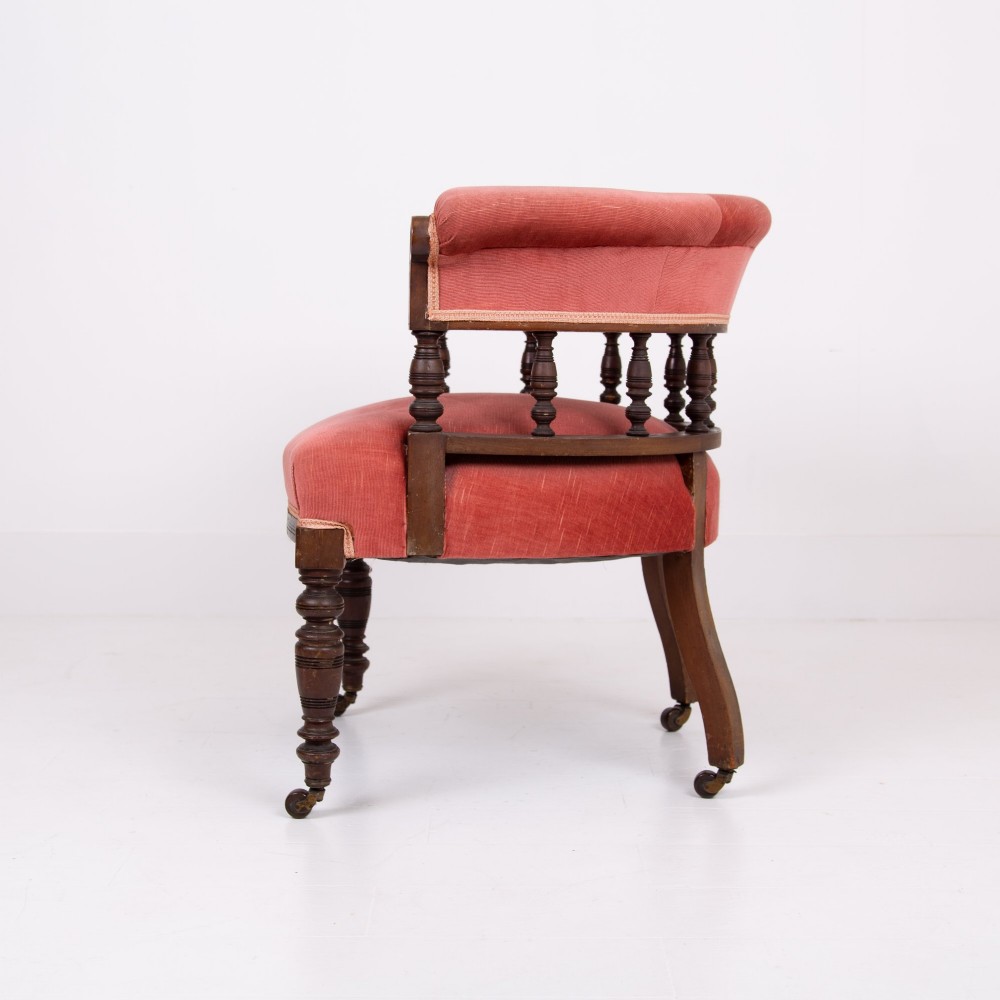 A late Victorian armchair featuring a curved horseshoe back rest, turned wood spindles, terracotta fabric upholstery, and turned front legs on castors. View II